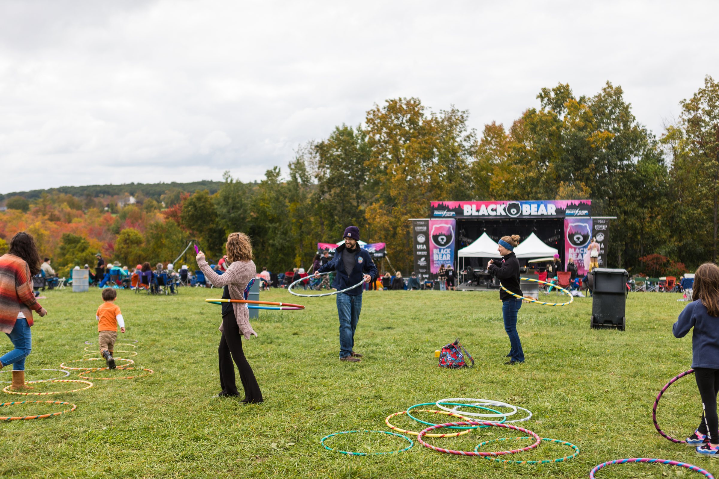 Families and children hula hooping on the open field at Black Bear Music Fest, with the stage and autumn treeline behind