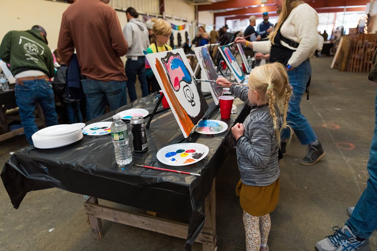 A child painting a Black Bear canvas at a long table during an art workshop