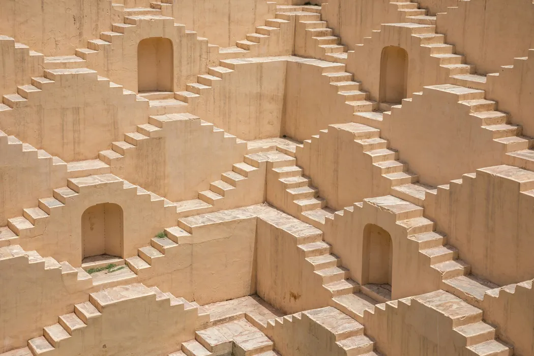 Symmetrical pattern of beige stone steps and staircases forming geometric shapes with small archways, part of a historic stepwell or architecture.
