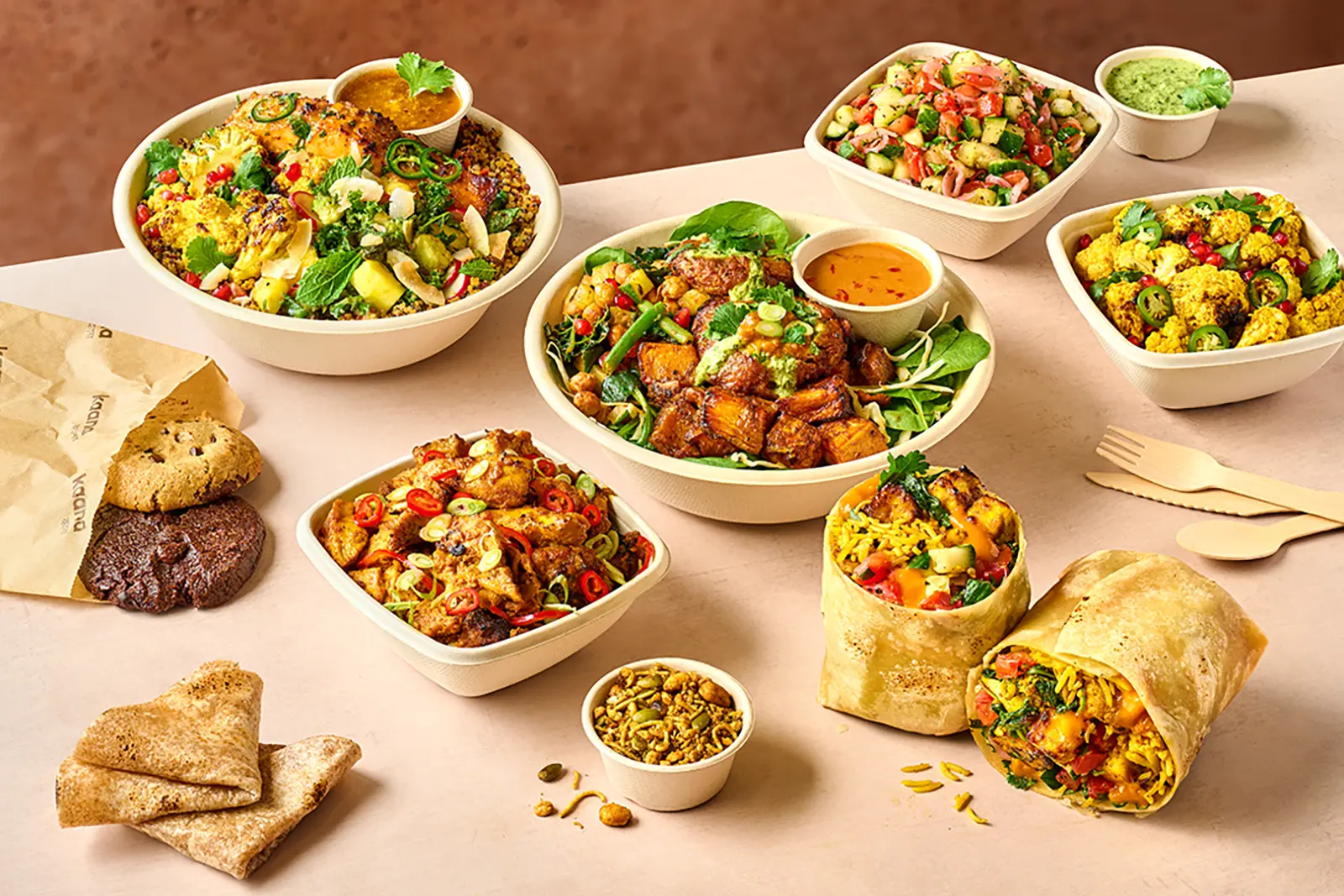 Assorted Indian-inspired meal with two bowls of roasted cauliflower with pomegranate seeds and green chili slices, a large bowl with roasted vegetables and green sauce with a small cup of dipping sauce, a square bowl with diced cucumber and tomato salad next to green chutney, a square bowl of spicy fried chicken pieces garnished with sliced red and green chili, two wraps filled with rice, vegetables, and paneer, a small cup of mixed nuts and seeds, chocolate chip and chocolate cookies partially inside a paper bag, and triangular folded flatbread on a light surface.