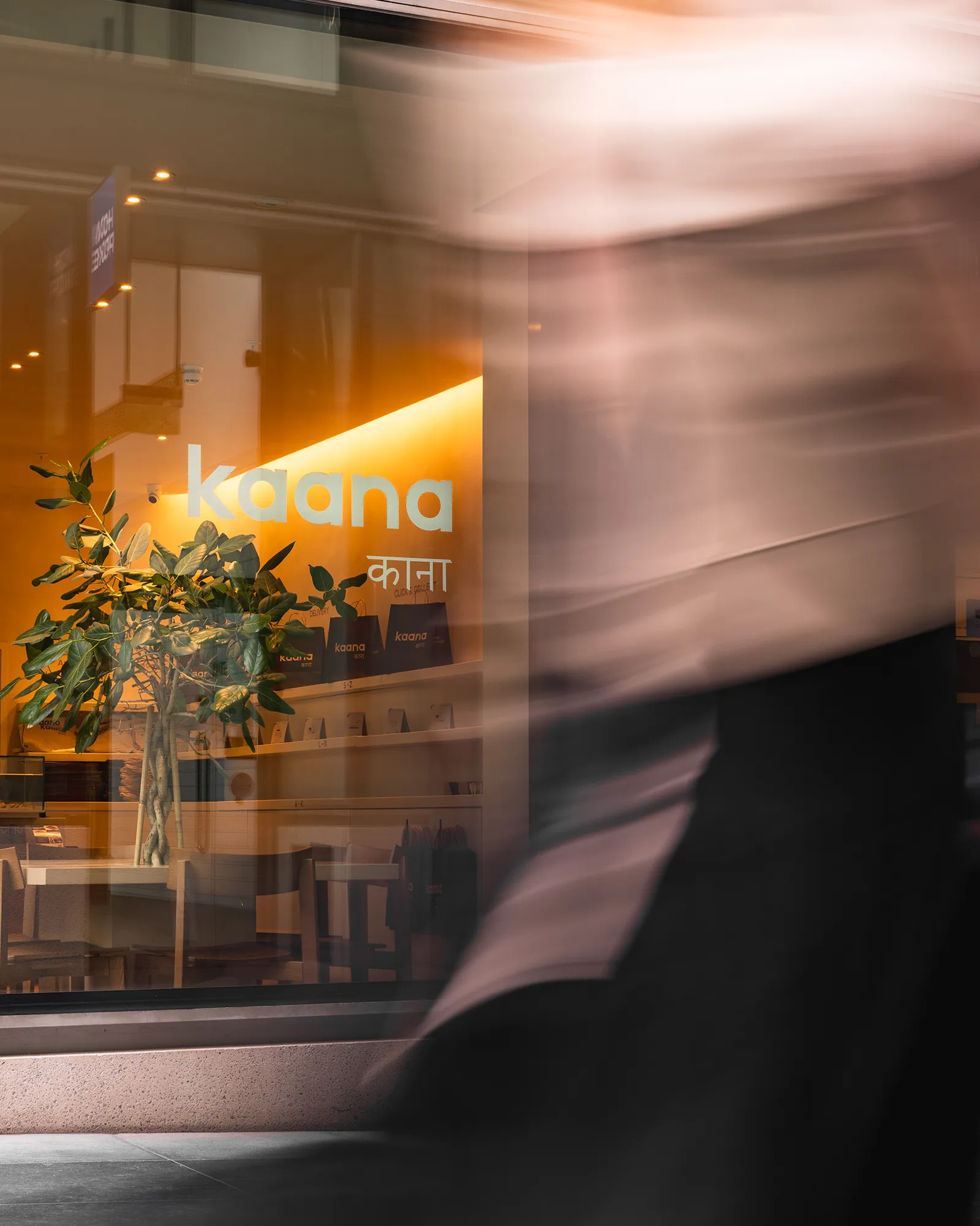 Kaana’s shopfront glowing warm at dusk, with a passer-by blurred in motion outside