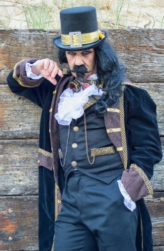 Man in steampunk style outfit with a brass coloured band around a top hat, airforce blue trousers and low-cut waistcoat, a white frill dress shirt and purple coat with gold brocade.