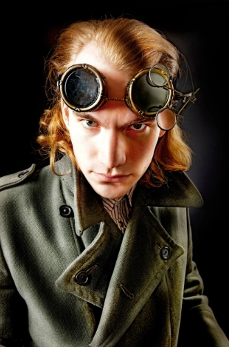Man in steampunk style outfit with goggles around his head, and a dark green or khaki trench coat