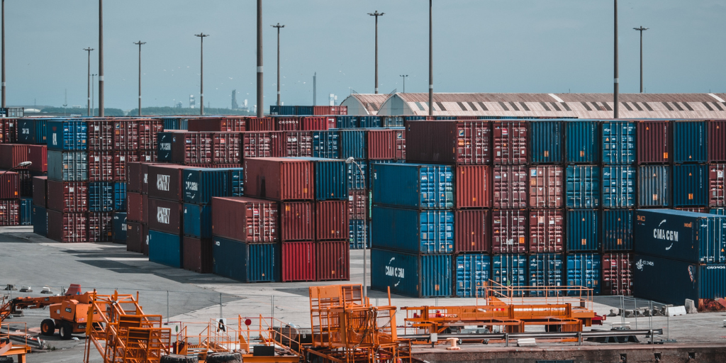 Aerial view of a busy container port with stacked shipping containers in blue and red colors, alongside orange cargo handling equipment, illustrating global supply chain logistics and freight operations