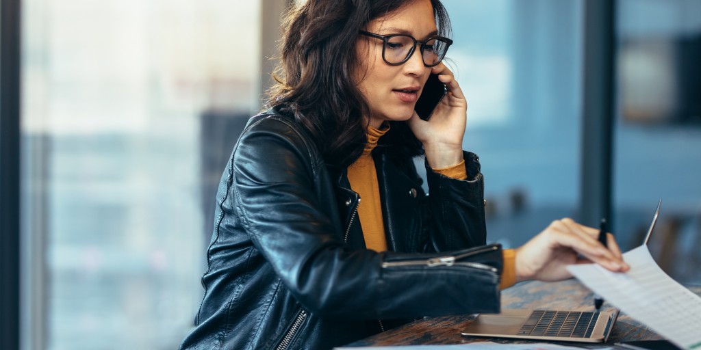 Professional data analyst in a modern office reviewing documents and talking on a smartphone while working on a laptop.