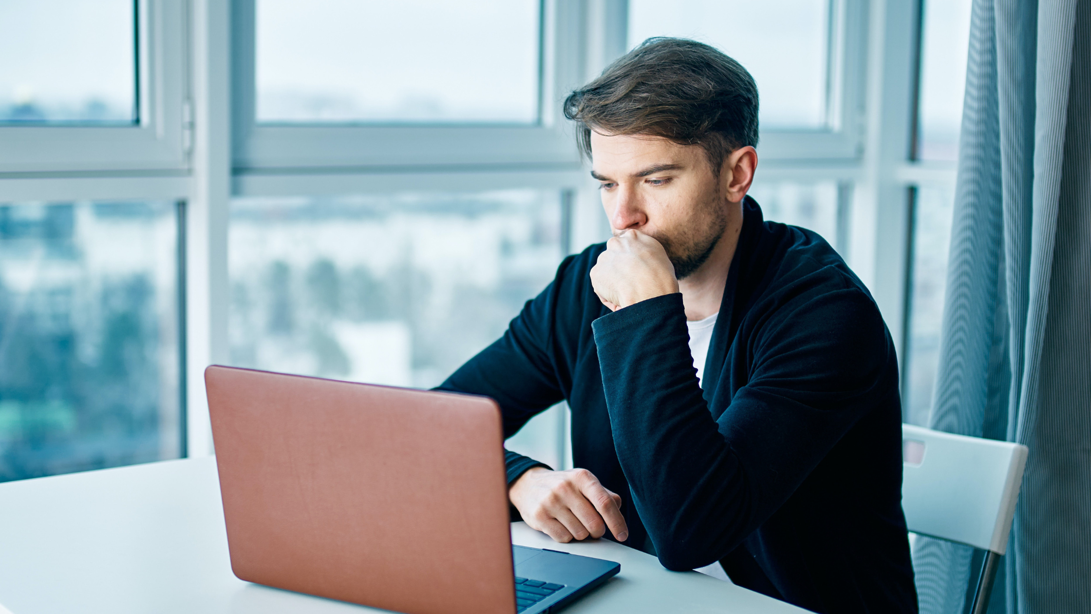 Man working at a laptop by a window, reading with his hand to his mouth