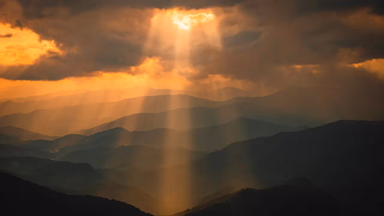 Shafts of golden sunlight pour through a gap in dark storm clouds, lighting up layer after layer of hazy mountain ridges below.