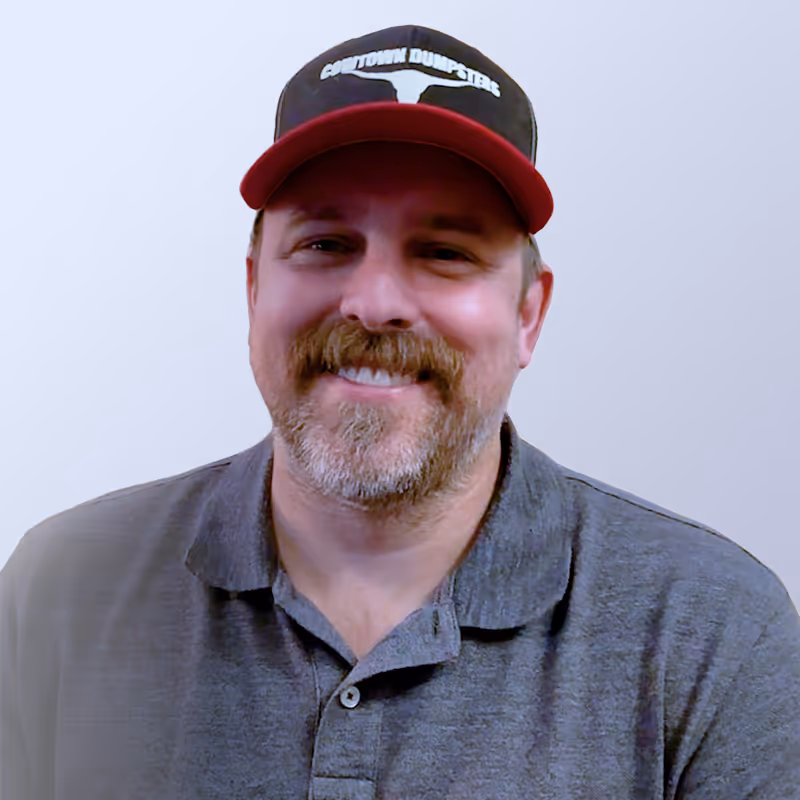 a smiling white man that is putting on a black face cap with the cowtown dumpsters inscription.