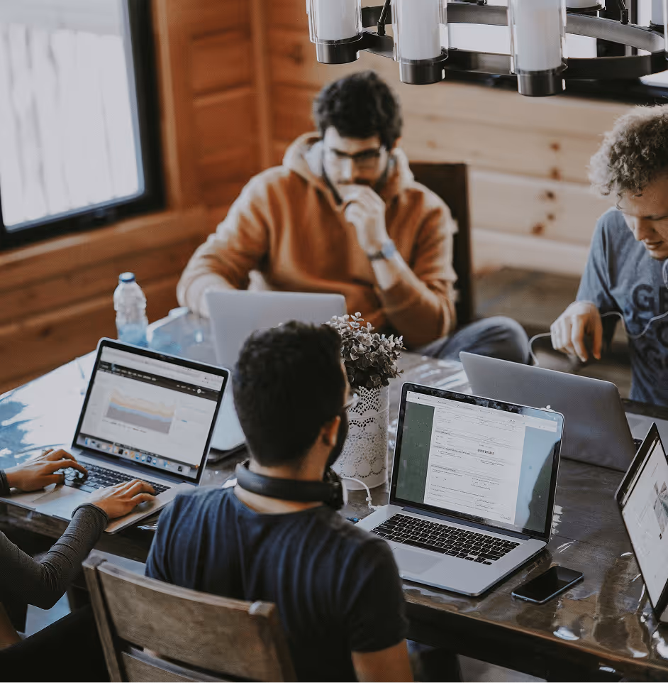 A group of four young people working on laptops around a wooden table in a cozy room with natural light.