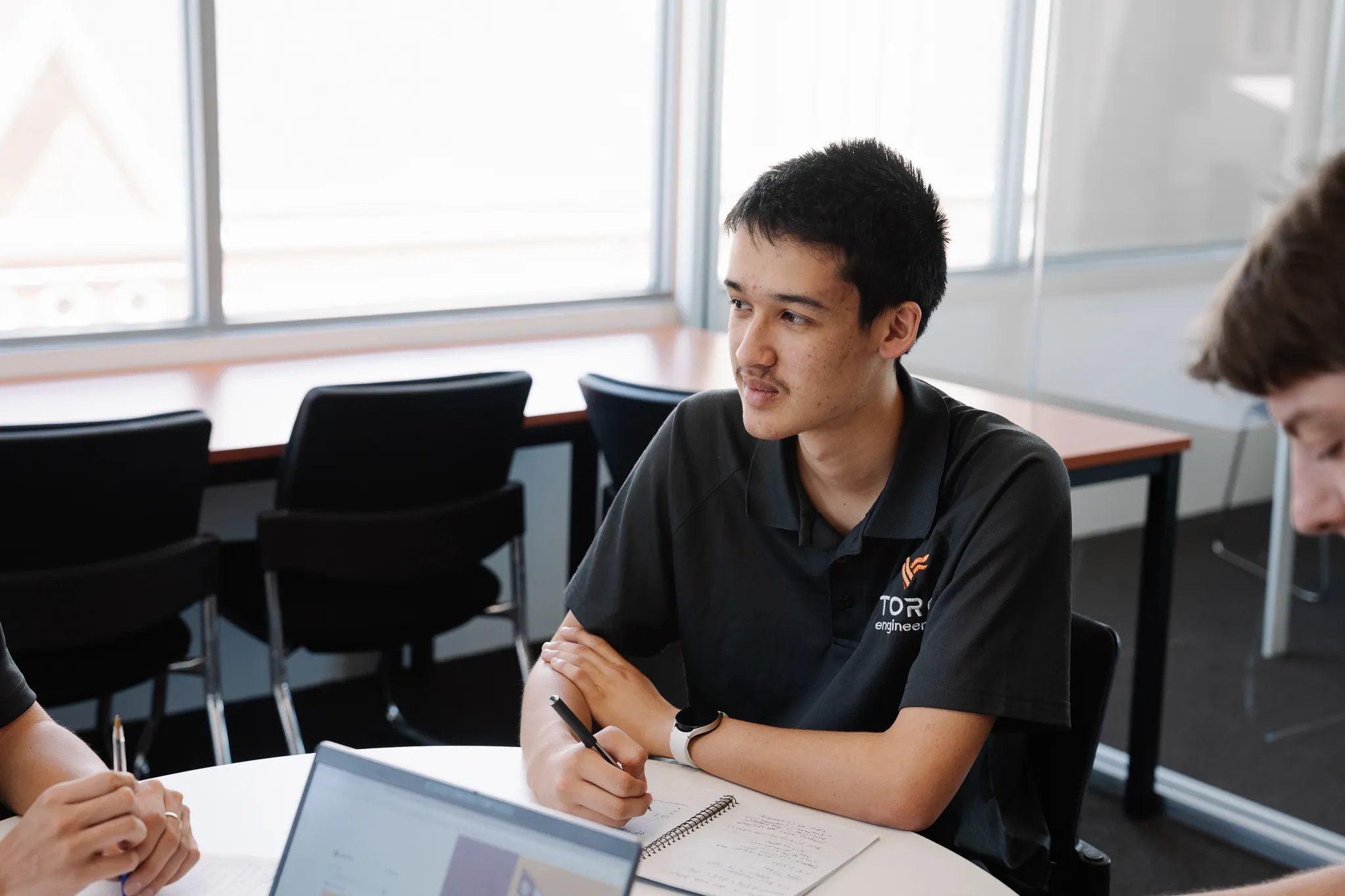 Young man in a black polo shirt with Toro Engineering logo sitting at a round table writing in a notebook.