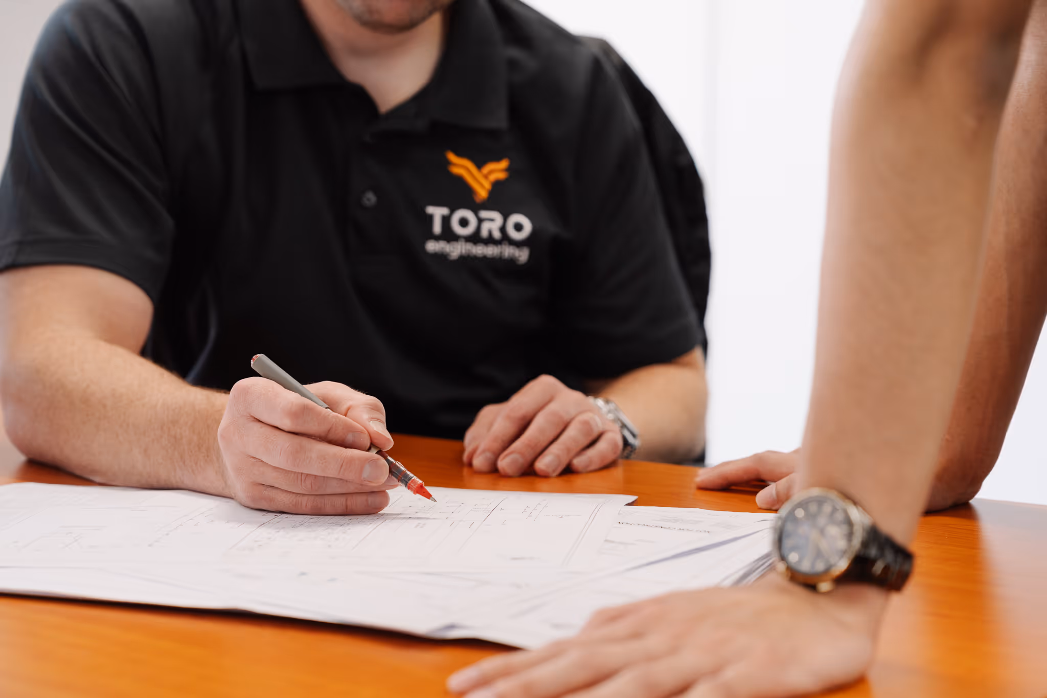 Person in a black Toro Engineering shirt marking architectural blueprints on a wooden table with a red pen while another person leans on the table.