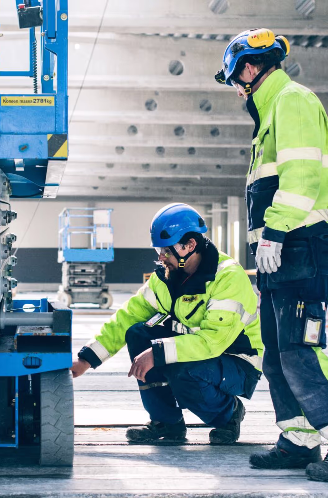 Two construction workers in high-visibility yellow jackets and safety helmets inspecting a blue Ramirent aerial lift platform. One worker is kneeling to examine the base of the equipment while the other stands watching.