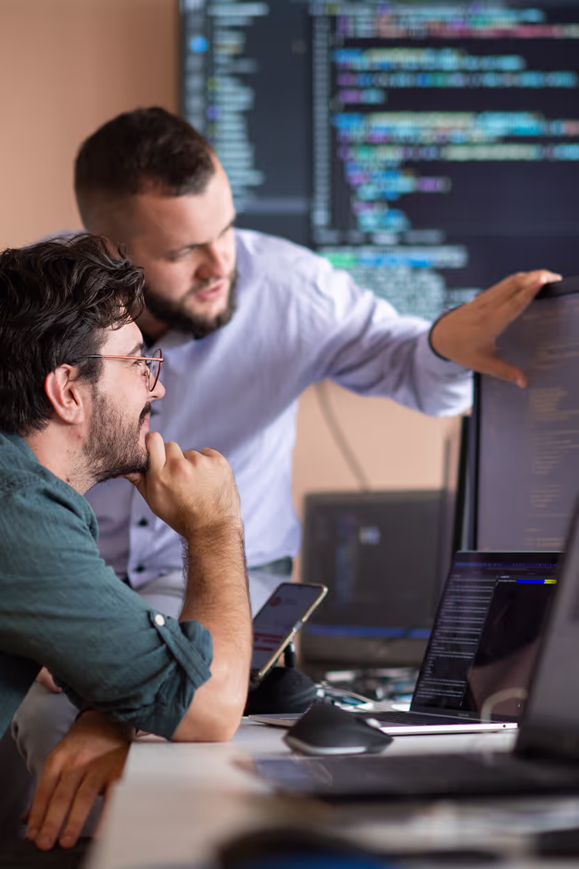 Two men are collaborating in front of a computer screen displaying code, with one pointing at the screen and the other thoughtfully engaged.