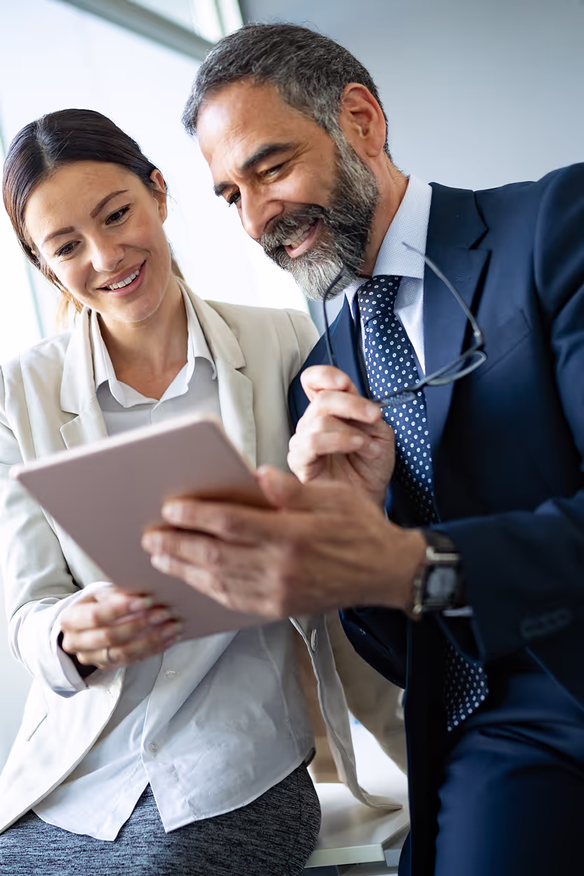 A man in a suit and a woman in business attire are smiling while looking at a tablet, suggesting collaboration or a business discussion.