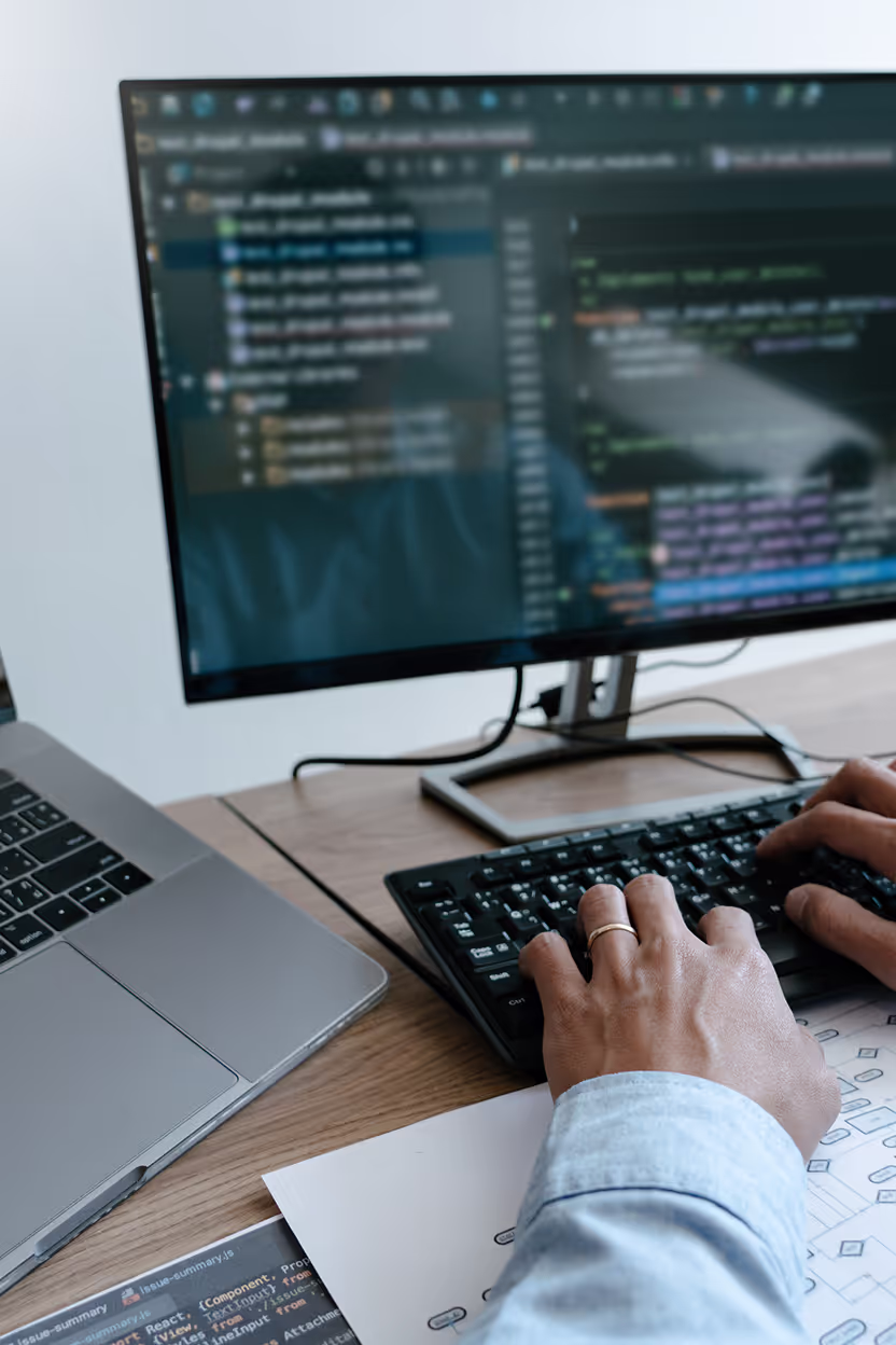 A close-up of hands typing on a keyboard, with a computer screen displaying code and a laptop on the desk, illustrating software development or programming.