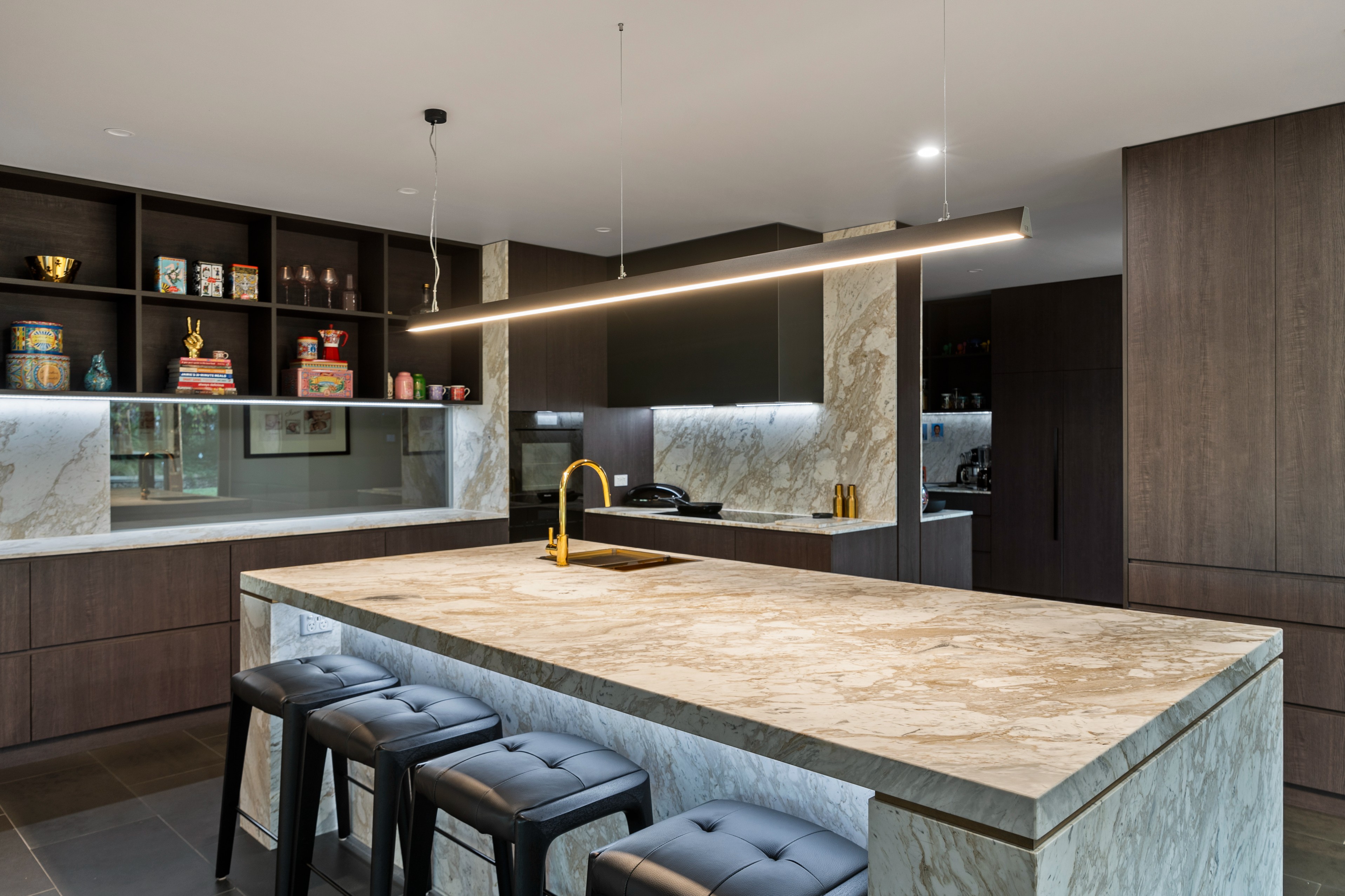 Modern kitchen with marble island countertop, gold faucet, black cushioned stools, and dark wood cabinetry with open shelves.