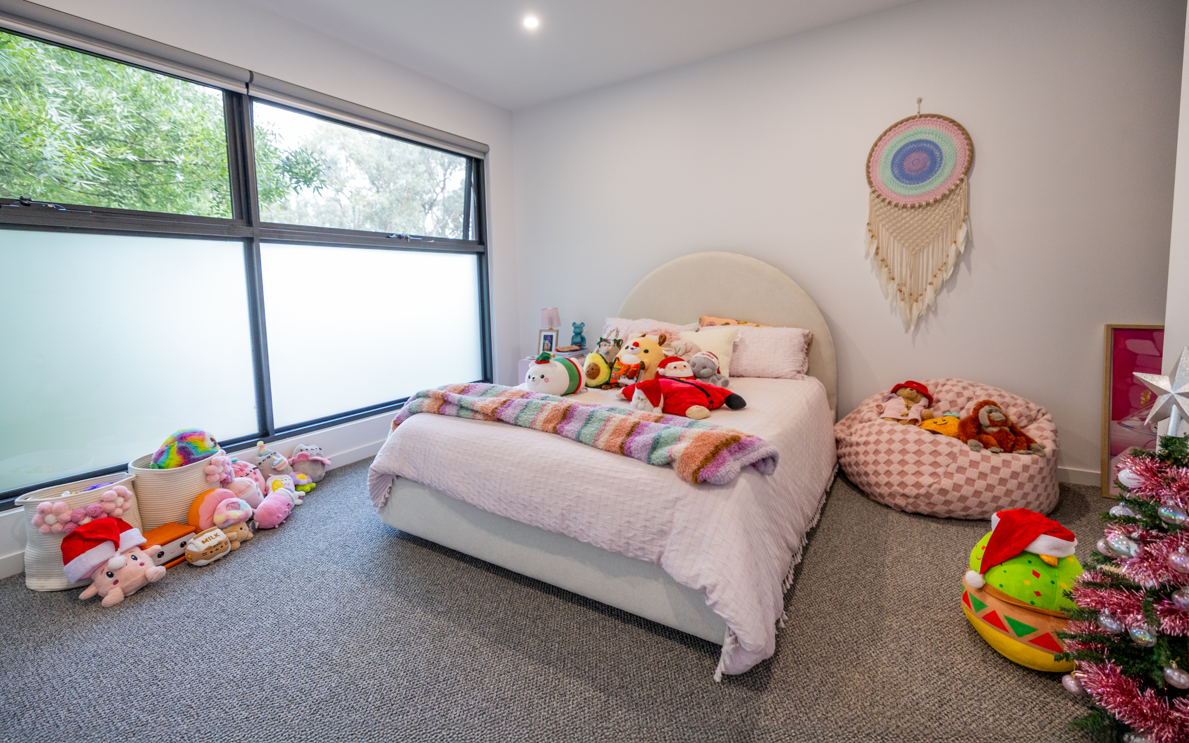 Cozy bedroom with a bed covered in a light pink quilt, colorful plush toys on the bed, large windows on the left, and a pink bean bag filled with stuffed animals.