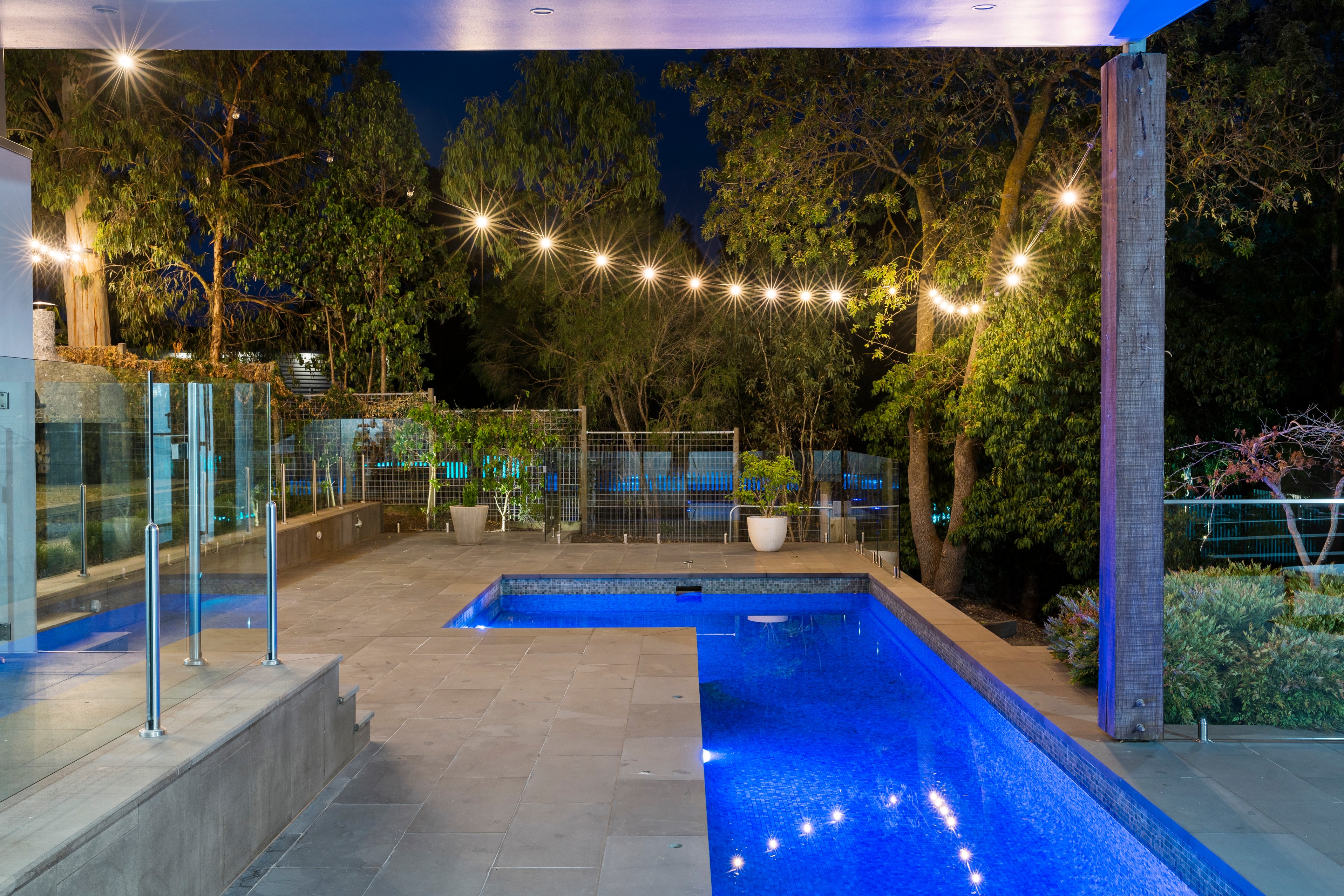 Modern outdoor swimming pool at night surrounded by glass fencing and string lights with trees in the background.