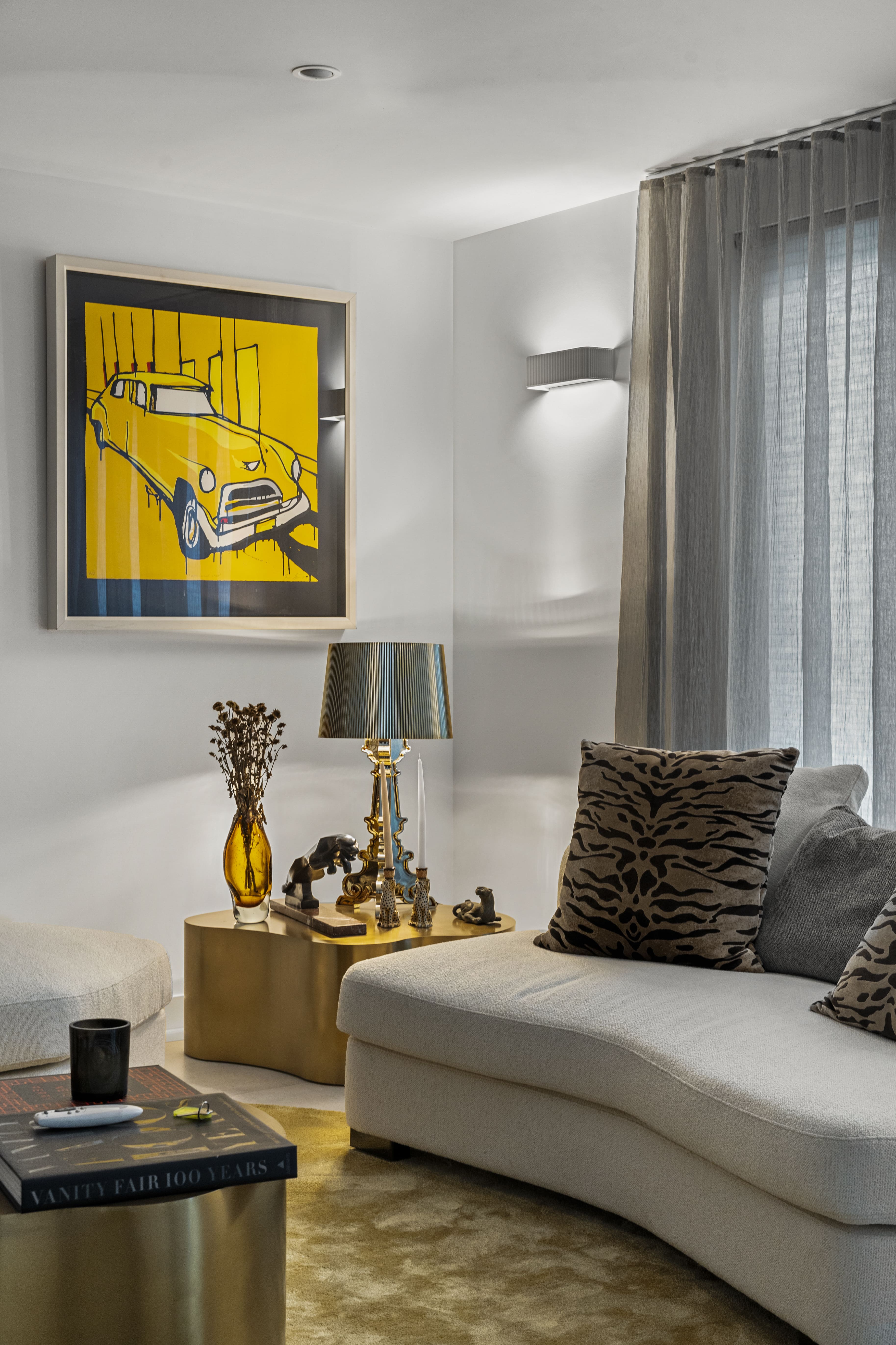 Modern living room corner with a white sofa, animal print pillows, gold side tables, a lamp, and a framed yellow car artwork on the wall.