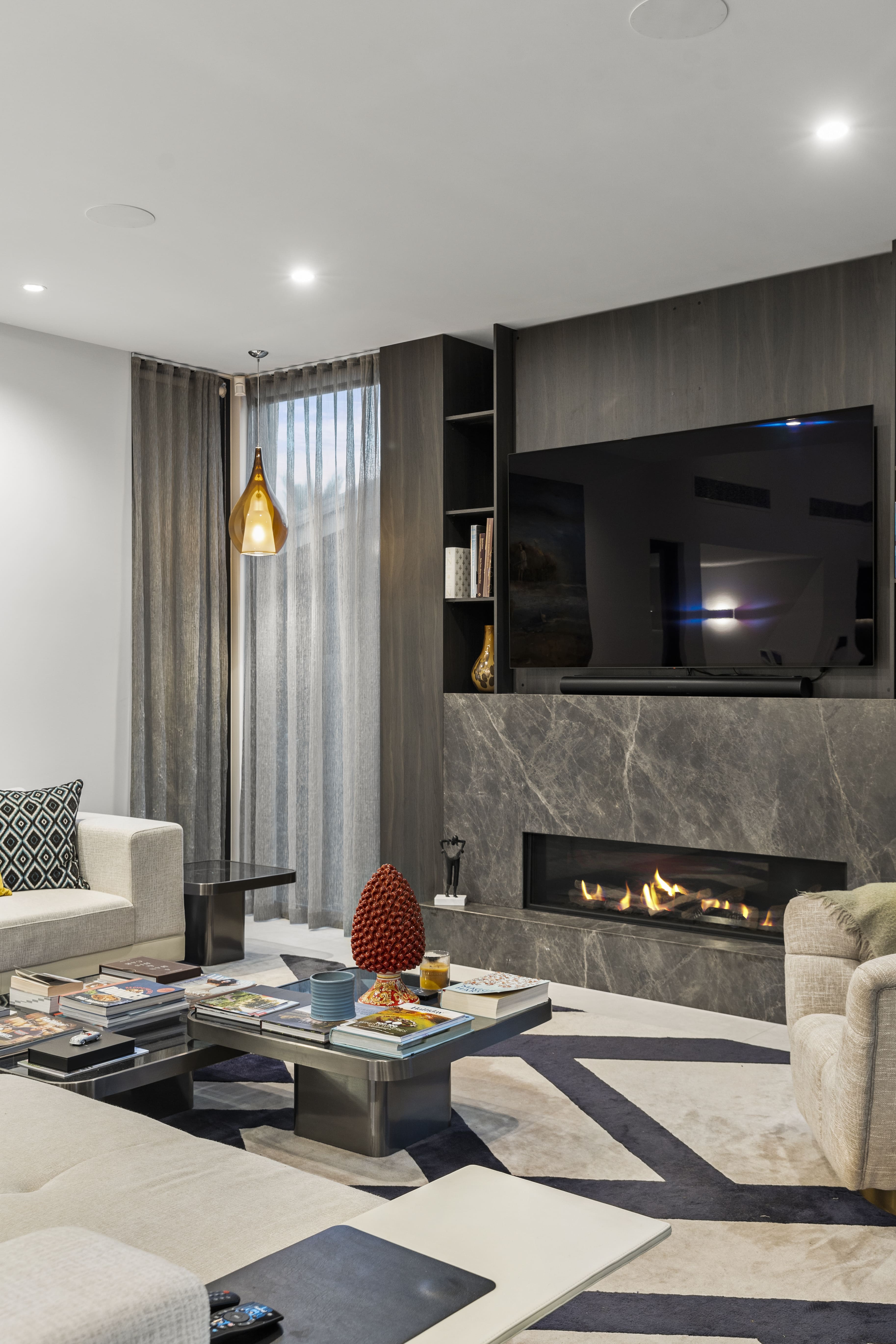 Modern living room with beige sofas, a coffee table stacked with books, a lit fireplace beneath a mounted flat-screen TV, and sheer curtains with a hanging amber pendant light.