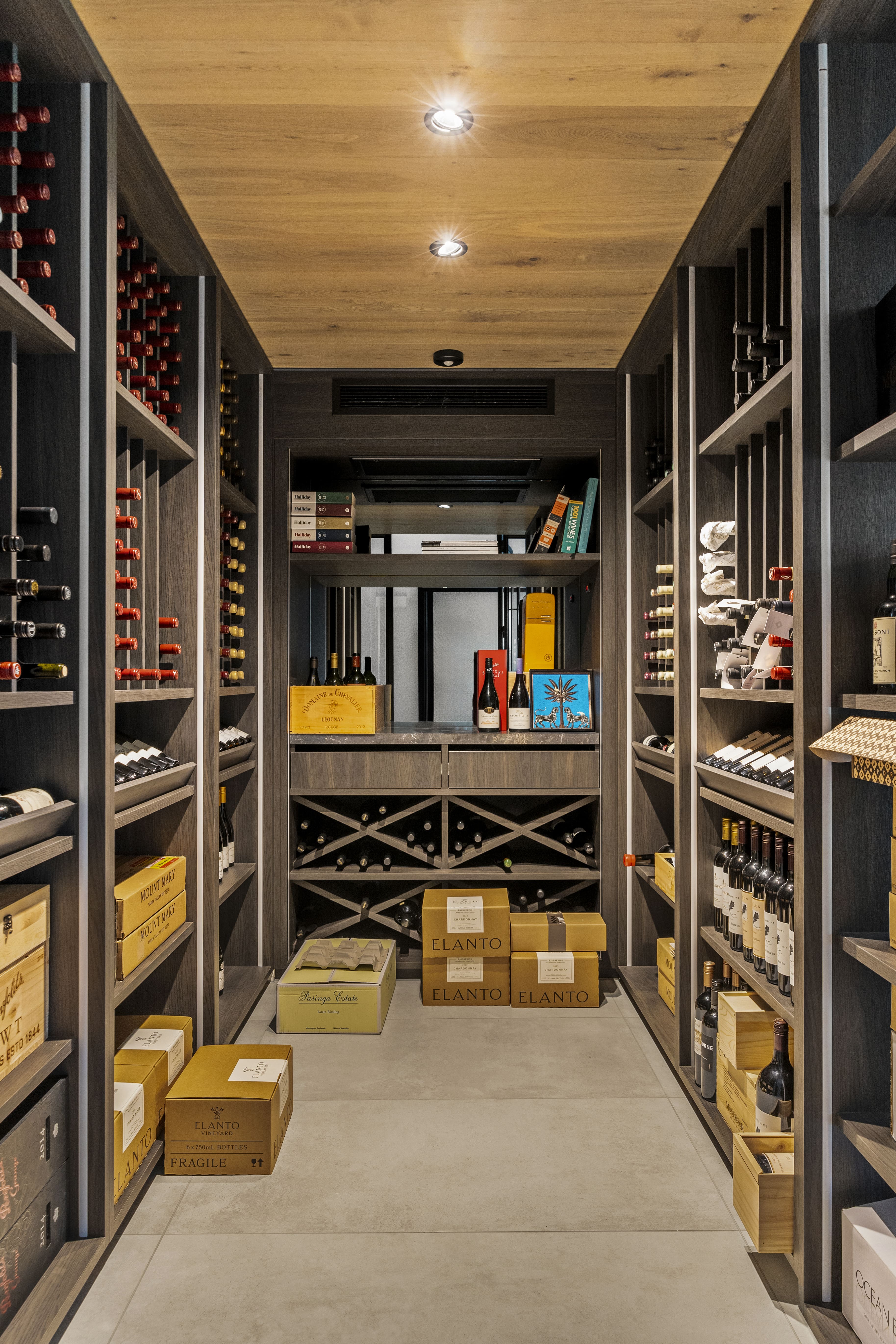 Modern wine cellar with wooden shelves filled with wine bottles, boxes on the floor, and a wooden ceiling with recessed lights.