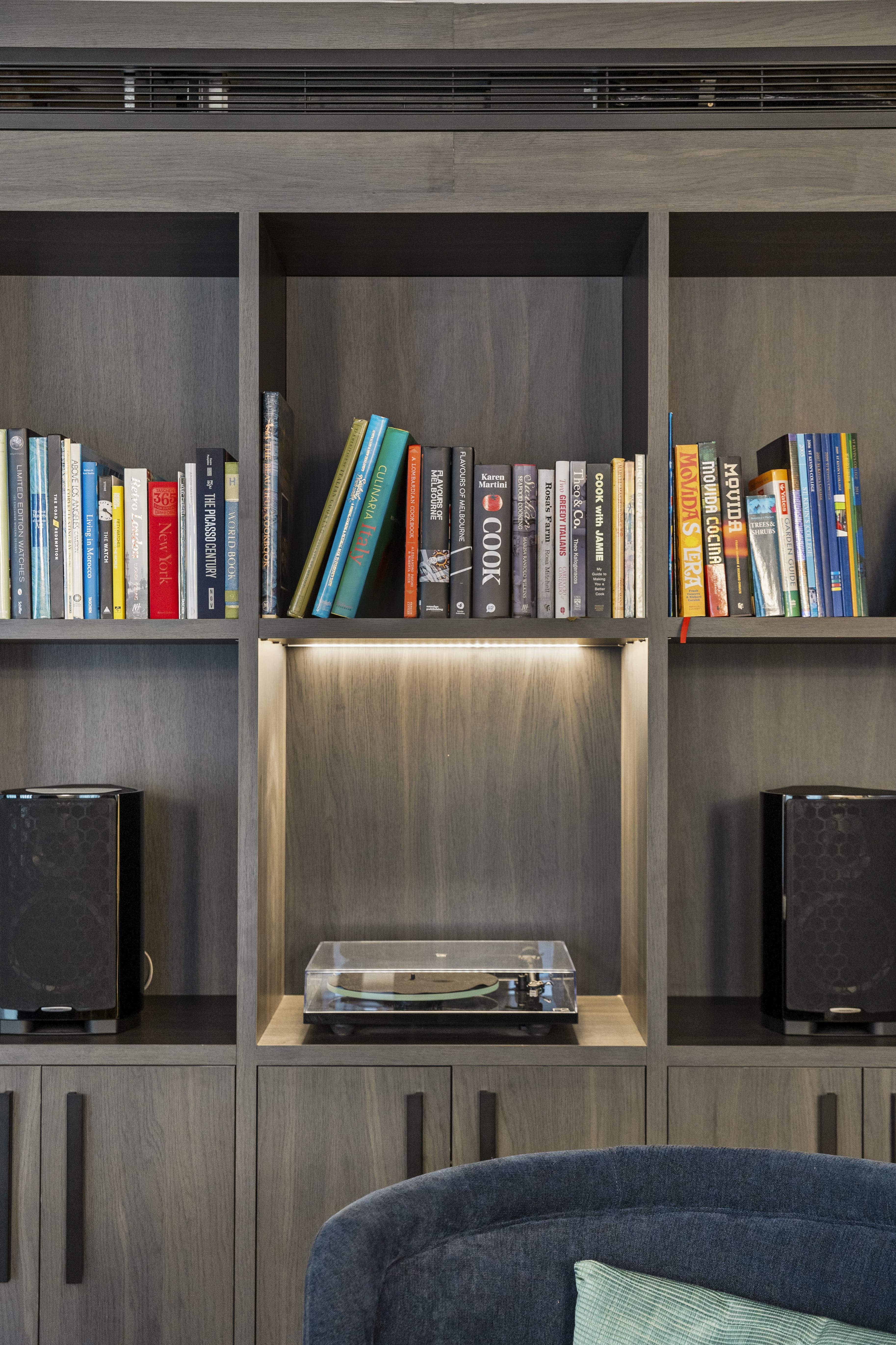 Wooden bookshelf with assorted books on top shelves, two black speakers on side shelves, and a turntable with a clear cover in the center cubby lit by an overhead light.