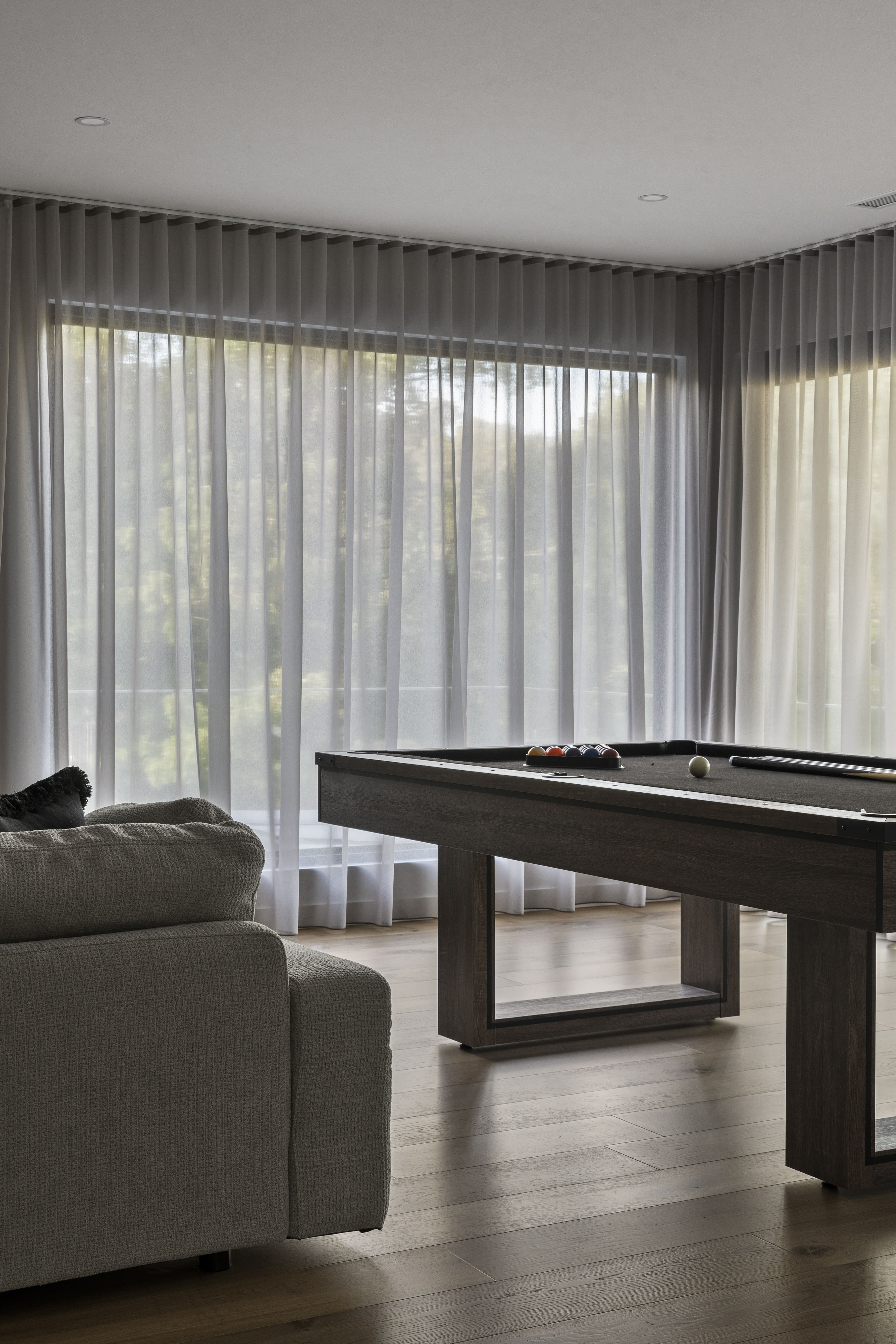 Modern room with sheer curtains, a gray textured sofa, and a dark wood pool table with balls and a cue stick on it.