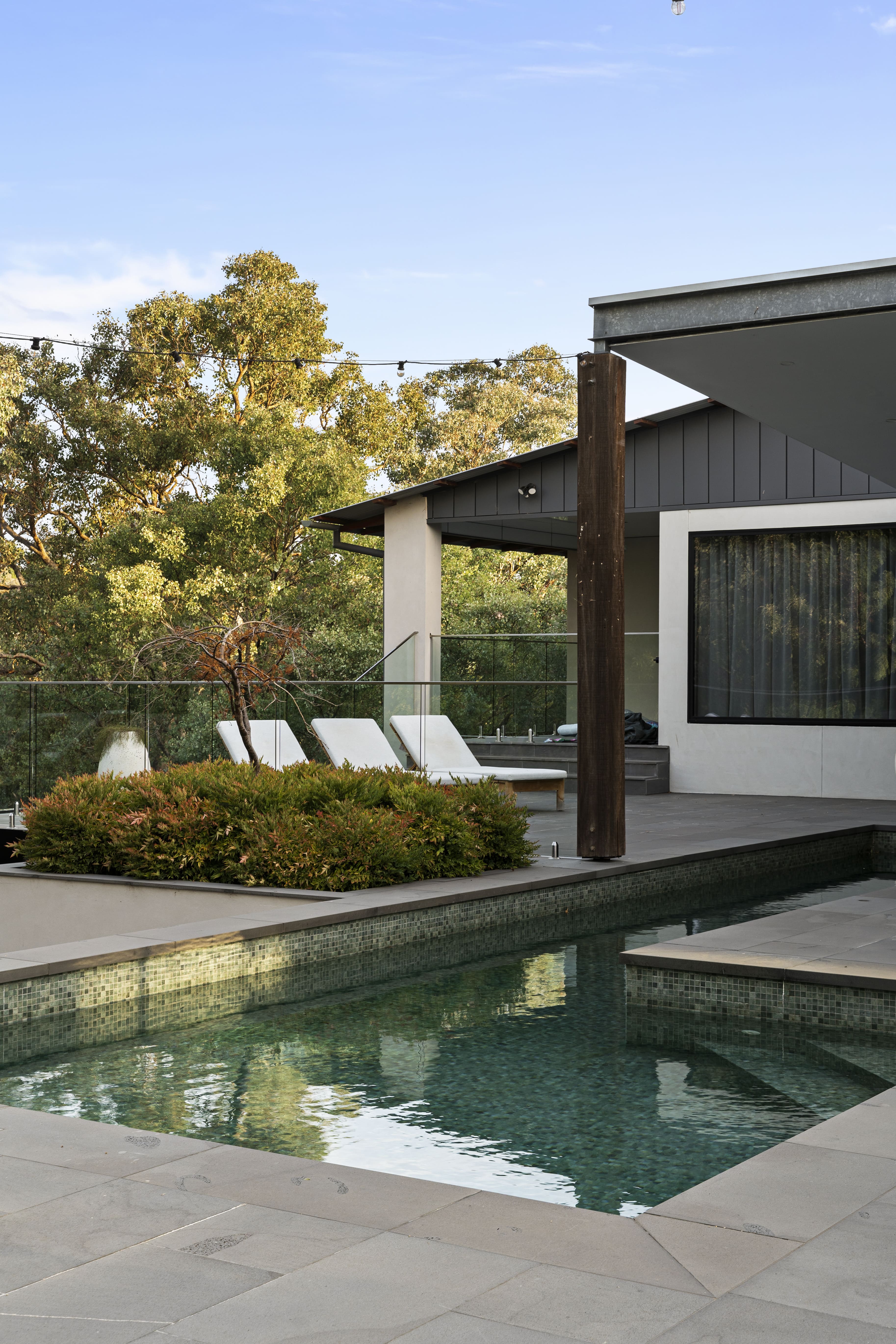 Modern outdoor pool area with clear water, surrounded by stone tiles, lounge chairs, and greenery under a blue sky.
