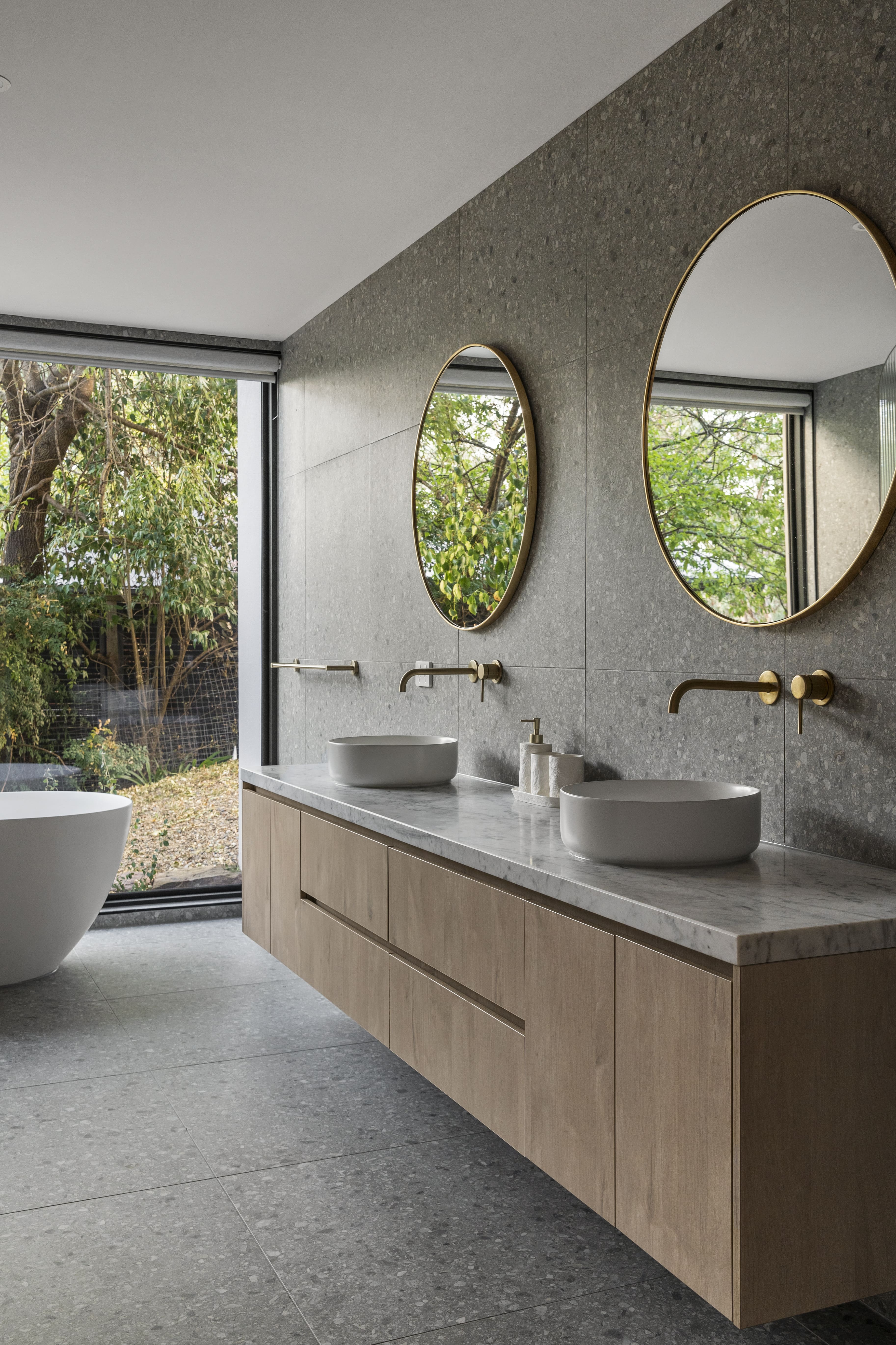 Modern bathroom with dual round mirrors, vessel sinks, gold faucets, and a large window showing green trees.
