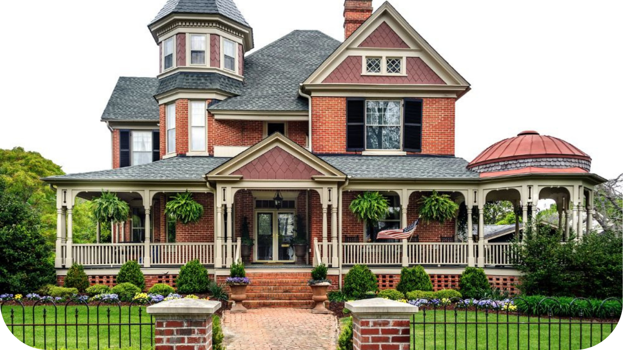 Victorian-style red brick home with ornate detailing, wraparound veranda, pitched roof, and lush landscaped front garden.