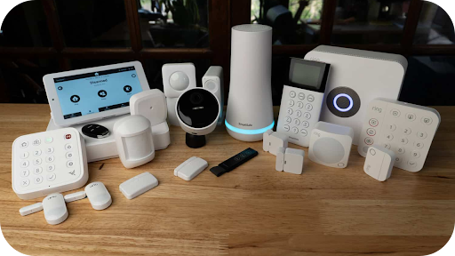 Assorted smart home security devices on a wooden table, including cameras, keypads, sensors, and a touchscreen control panel.