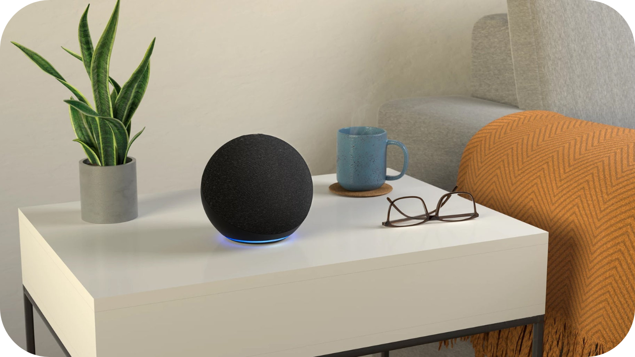 A modern living room setup with a black smart speaker glowing blue on a white side table, next to a potted plant, blue ceramic mug on a coaster, and a pair of glasses by an orange throw on a grey sofa.