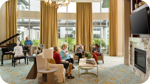 Seniors socialising in a bright lounge with tall golden curtains, large windows, piano, fireplace, and modern furnishings.