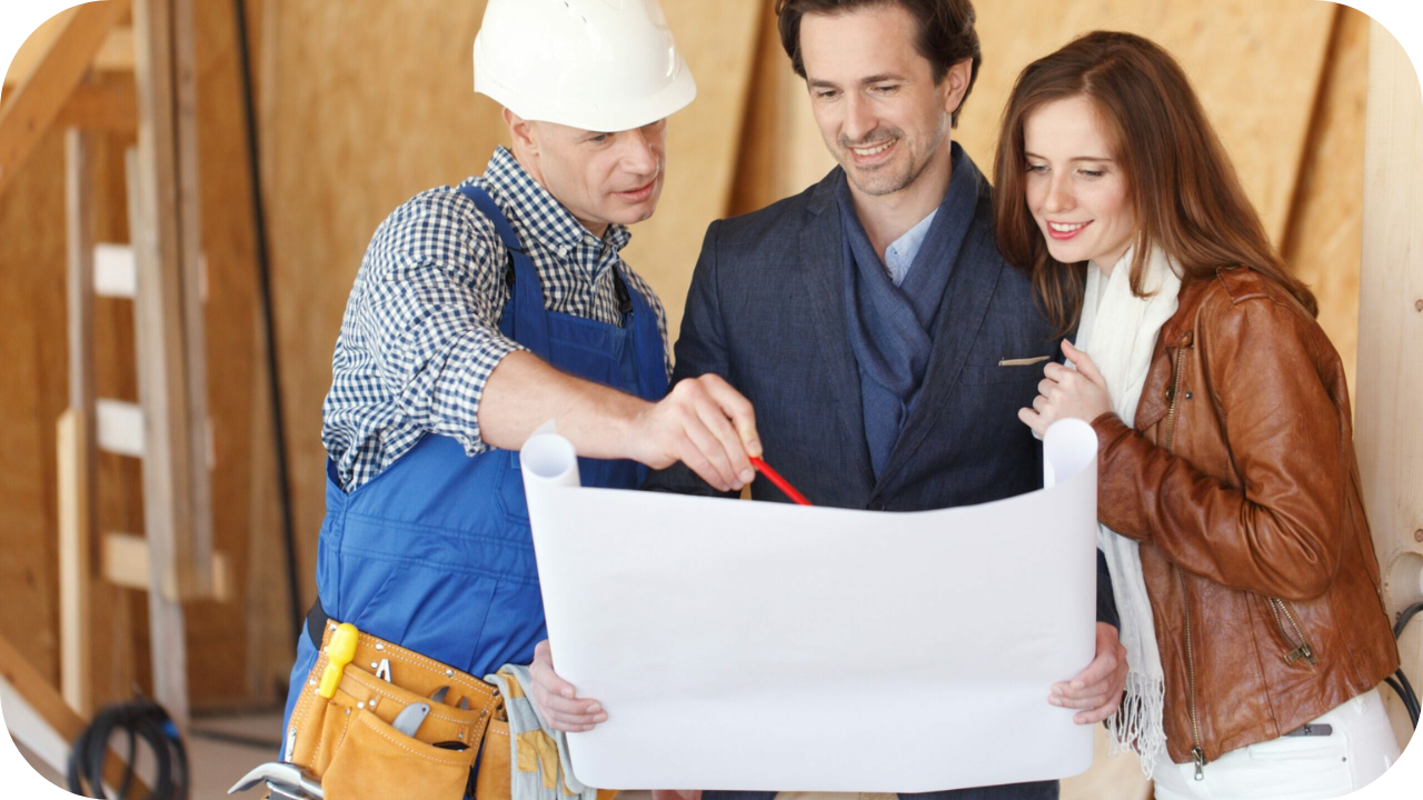 Builder in a hard hat showing house plans to a couple during construction planning inside an unfinished wooden frame.