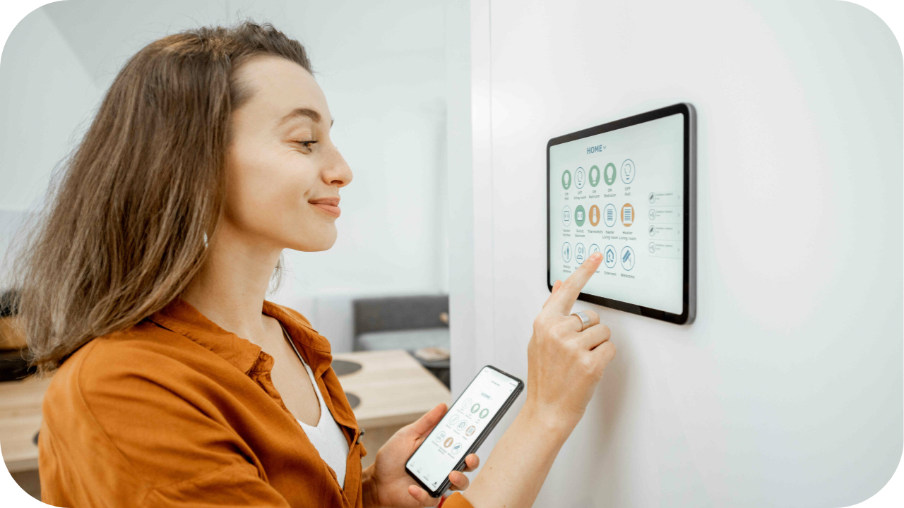 Woman using a wall-mounted smart home control panel while holding a smartphone, managing connected devices and home automation settings with a smile.