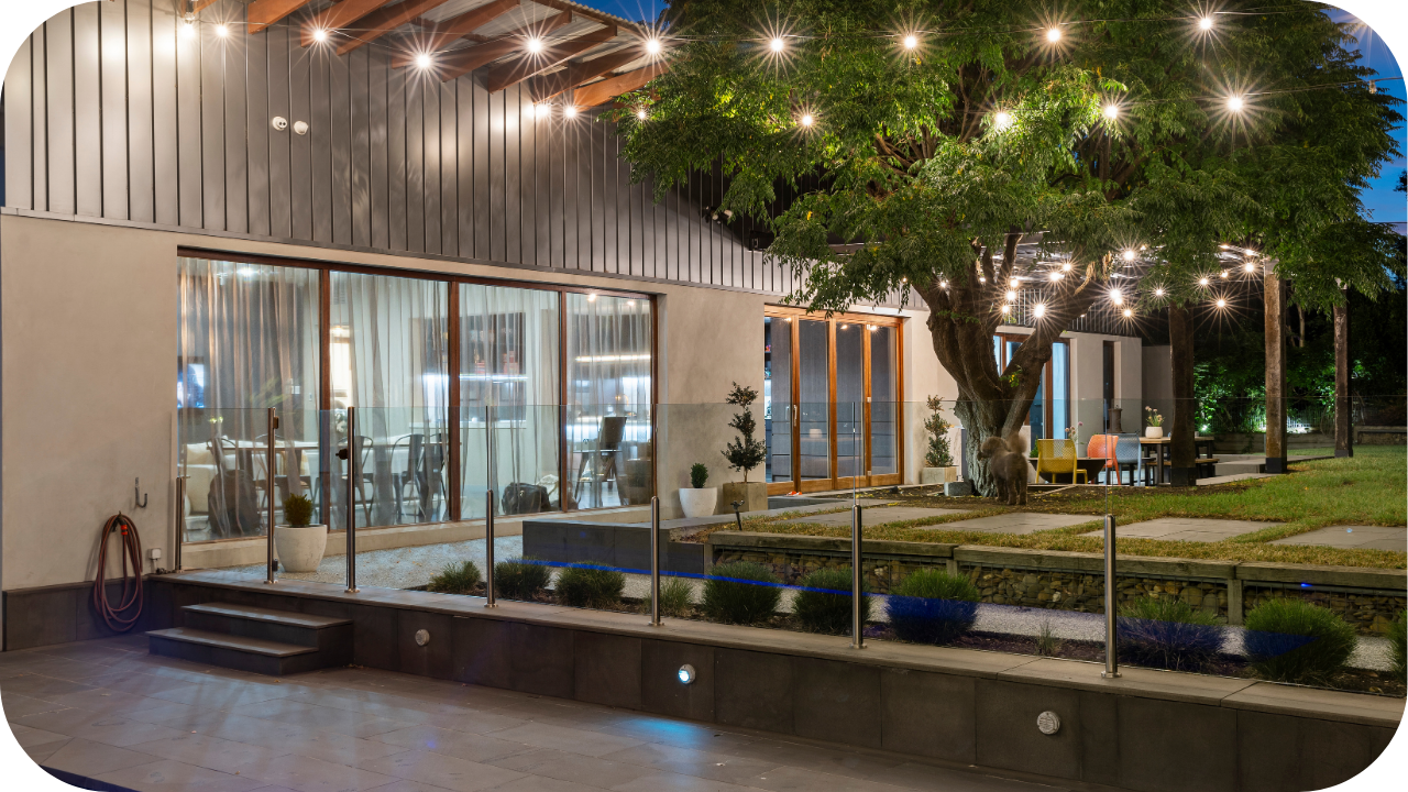 Modern outdoor entertaining area with string lights, glass fencing, a poolside view, and a large tree beside a dining table set under the illuminated night sky.