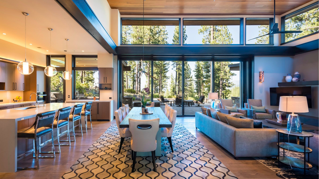 Spacious open plan living, dining, and kitchen area with full height windows showcasing forest views and warm modern interior styling.