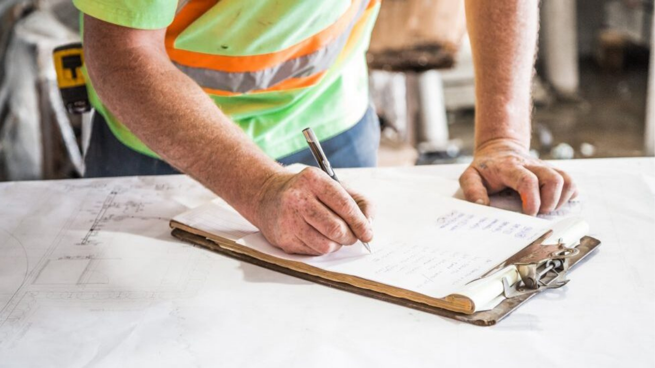Builder reviewing notes on a clipboard while preparing construction plans on a site table during early pre-construction planning