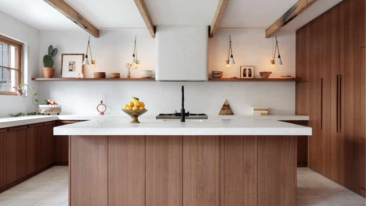Warm modern kitchen with timber cabinetry, white stone benchtops, open shelves, pendant lighting and exposed ceiling beams styled with fruit and decor.