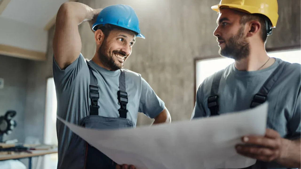 Two construction workers are reviewing building plans on site, wearing safety helmets and discussing project details with a relaxed, friendly tone.