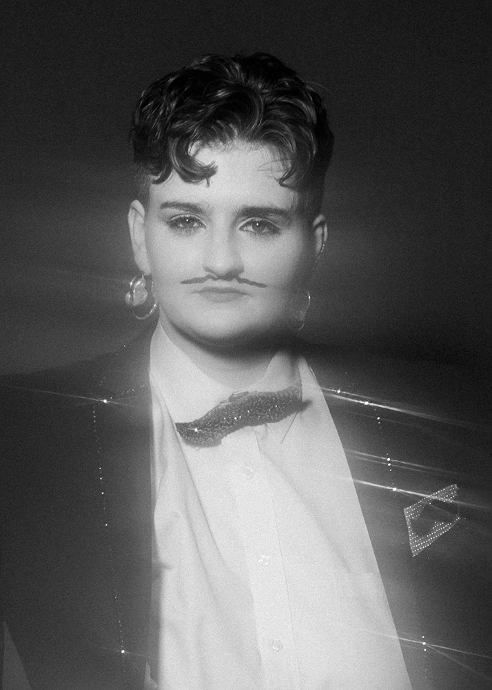 Black and white portrait of a person with short curly hair, wearing a blazer, white shirt, bow tie, hoop earrings, and a drawn-on mustache.