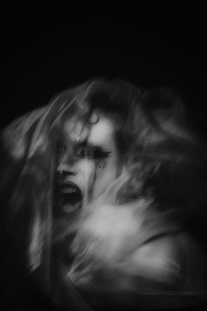Black and white blurred image of a person with dramatic makeup, mouth open as if screaming, partially obscured by translucent fabric.