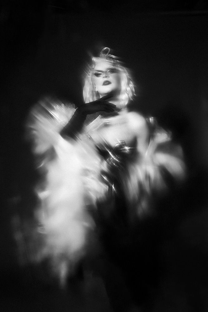 Black and white blurred portrait of a glamorous person wearing dark gloves and a feathered garment.