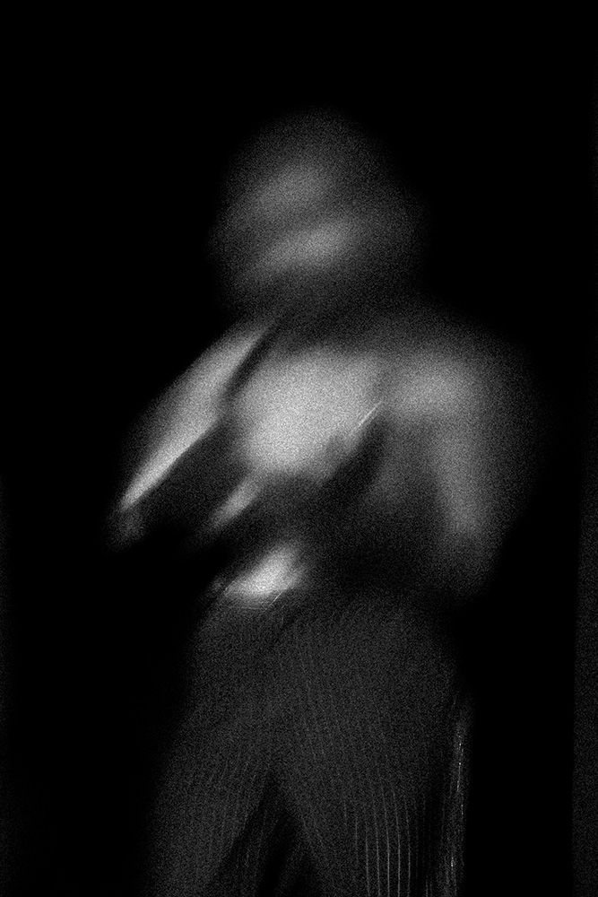 Black and white blurred image of a person with bare upper body and striped pants against a dark background.