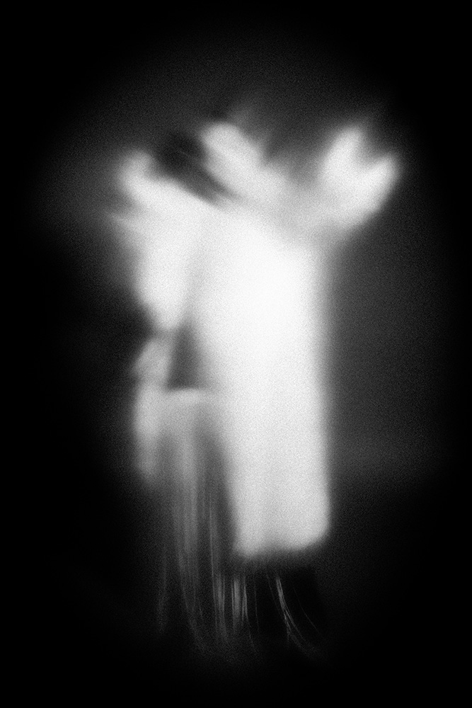 Abstract blurry black and white image suggesting two ghostly figures with outstretched arms against a dark background.