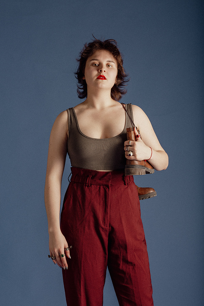 Young woman with short dark hair, wearing a gray crop top and high-waisted red pants, holding brown boots over her shoulder against a blue background.