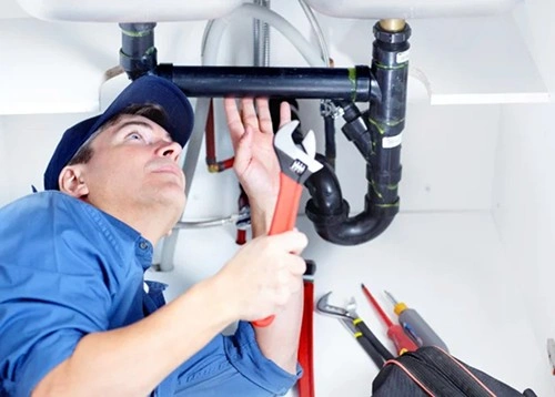 Plumber in blue uniform fixing pipes under sink with wrench