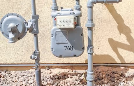 Gray utility meter with pipes mounted on stucco wall with rocky ground