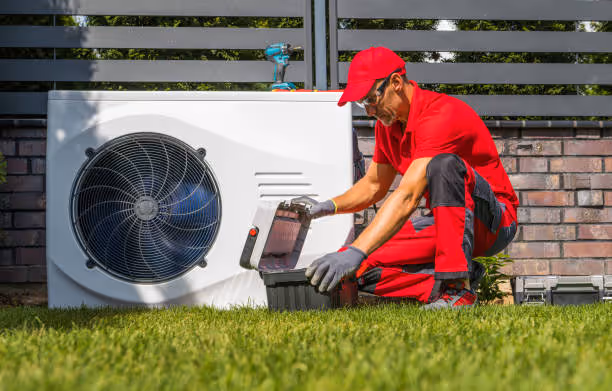 HVAC technician in red uniform servicing outdoor heat pump unit on grass