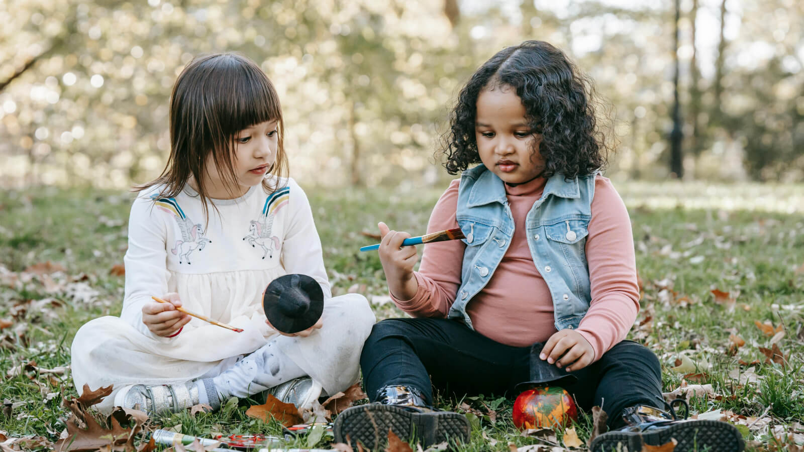 two little girls sit painting pumpkins in the park